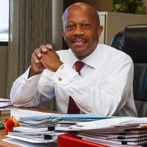 Professional portrait of Sanford City Manager Norton Bonaparte Jr. sitting at his office desk, looking directly at the camera with a warm smile. Key visual details include: Subject: City Manager Norton Bonaparte Jr., wearing a white button-down dress shirt with French cuffs, cuff links, and a red pattern tie, resting his clasped hands on the desk. Setting: An office environment with stacks of organized documents, folders, and binder-clipped papers neatly arranged on the wooden desk in front of him. A black executive office chair and indoor houseplant are visible in the background.