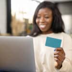 Cheerful young multiracial woman shopping online with credit card and laptop while sitting on sofa at home,