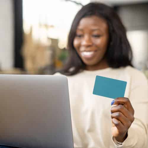 Cheerful young multiracial woman shopping online with credit card and laptop while sitting on sofa at home,