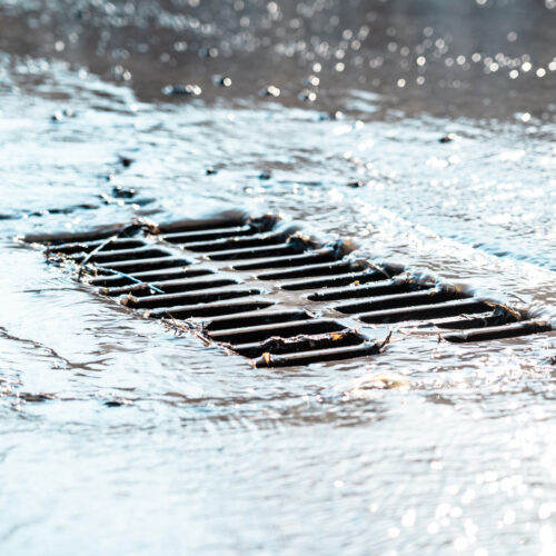 he grate of the storm sewer after the rain. The water drains into the storm drain. Sun glare, defocused background