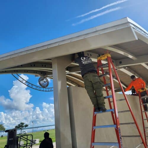 Two construction workers on ladders installing structural elements under the roof of an open-air park pavilion shelter overlooking water on a bright, sunny day. A decorative arch sign reading "Mellon Park" is visible in the background frame.