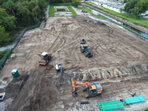High-angle aerial view of a cleared dirt construction site. Heavy machinery is scattered across the dirt lot, including an orange Doosan tracked excavator in the foreground, an orange front loader, an orange road roller, and a white mini-excavator. Green construction pipes lie bundled at the bottom right, while a drainage canal runs alongside the site near surrounding trees and industrial buildings in the background.