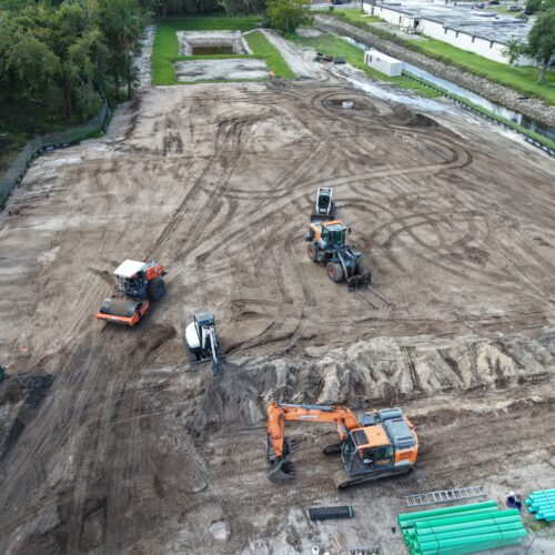 High-angle aerial view of a cleared dirt construction site. Heavy machinery is scattered across the dirt lot, including an orange Doosan tracked excavator in the foreground, an orange front loader, an orange road roller, and a white mini-excavator. Green construction pipes lie bundled at the bottom right, while a drainage canal runs alongside the site near surrounding trees and industrial buildings in the background.