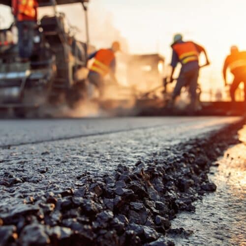A low-angle, close-up shot of freshly laid, dark asphalt with a rough aggregate texture in the foreground. In the blurred background, road construction crew members wearing high-visibility orange vests and hard hats work alongside heavy paving machinery at sunrise or sunset, backlit by warm golden light.